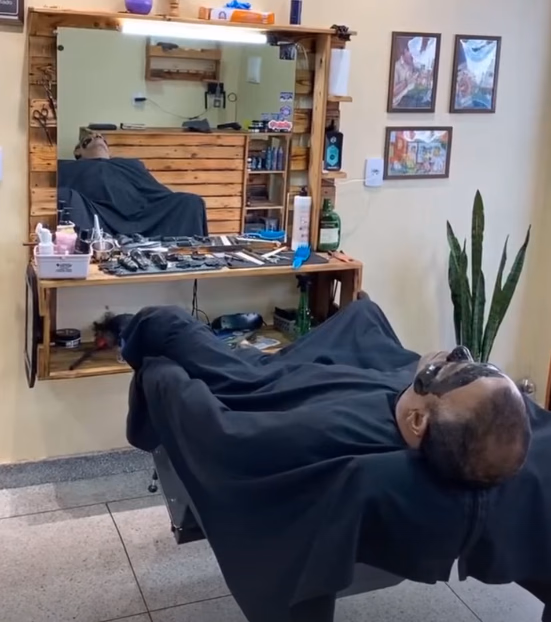 Interior da Lá Barbearia: bancada de madeira com espelho e cliente na cadeira reclinada durante limpeza de pele
